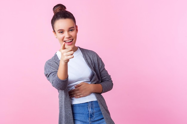 Oye, tú Retrato de una adolescente morena alegre con peinado de moño con ropa informal apuntando a la cámara sosteniendo su estómago y riéndose a carcajadas divirtiéndose con amigos en el estudio con fondo rosa