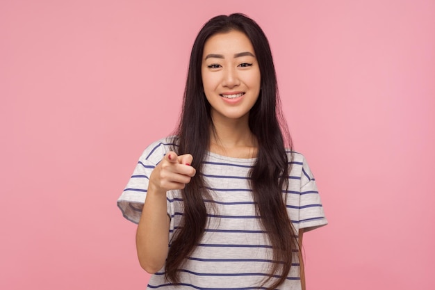 Oye, tú Retrato de una chica alegre con el pelo largo y moreno con una camiseta a rayas apuntando a la cámara y sonriendo gesticulando que necesitamos que elijas una foto de estudio interior aislada en un fondo rosa