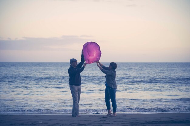 Paar am Strand fliegende Papierlaterne. Paar mittleren Alters mit chinesischer Laterne während des Sonnenuntergangs am Strand. Fröhliches Paar im Begriff, Papierlaterne auf Sand am malerischen Strand in die Luft zu entlassen?