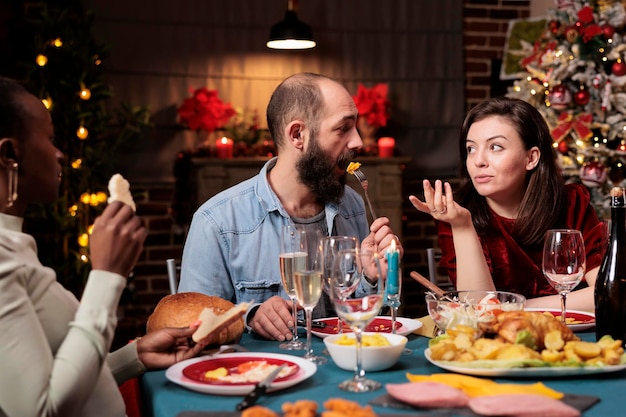 Foto paar, das weihnachtsabend mit der familie feiert, verhält sich romantisch am tisch mit menschen, verschiedene personen genießen das zusammenkommen mit hausgemachtem essen und alkohol, um die weihnachtsfeier zu feiern.