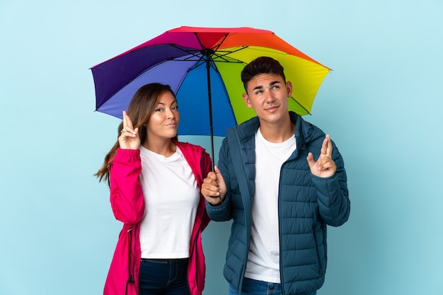 Paar hält einen Regenschirm auf Blau mit gekreuzten Fingern und wünscht das Beste