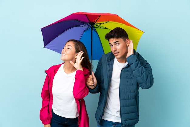 Paar hält einen Regenschirm auf Blau und hört etwas zu, indem es die Hand auf das Ohr legt