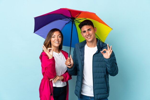 Paar hält einen Regenschirm auf Blau und zeigt ein OK-Zeichen mit den Fingern