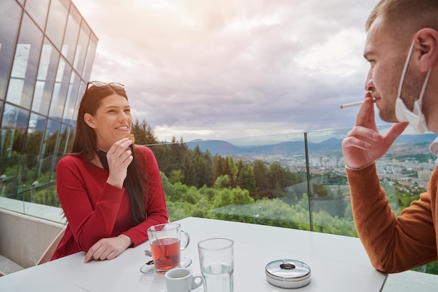 paar im restaurant mit medizinischen gesichtsschutzmasken coronavirus-schutz soziales distanzierungs- und präventionskonzept männliches rauchen und checken des smartphones