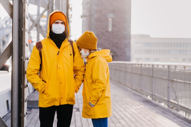 Paar in chirurgischen Gesichtsmasken in gelben Jacken, verwirrt wegen des Coronavirus, stehen auf der Brücke. Mädchen ist besorgt in der Schulter ihres Freundes begraben. Covid-19-Konzept.