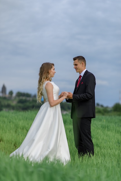 Paar in Hochzeitskleidung steht auf einer grünen Wiese auf dem Hintergrund des Dorfes bei Sonnenuntergang, die Braut und der Bräutigam