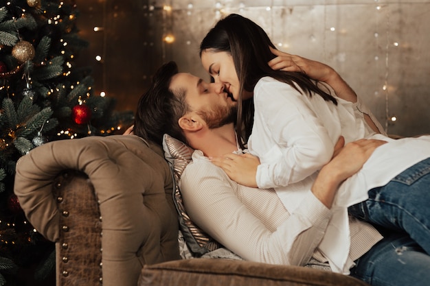 Paar in weißen Hemden umarmen und wollen auf der beige Couch nahe dem Weihnachtsbaum küssen.