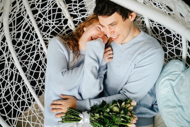 Paar Mädchen und Kerl umarmen sich auf einem großen Stuhl mit einem Strauß Rosen. Weißer und blauer Pullover.