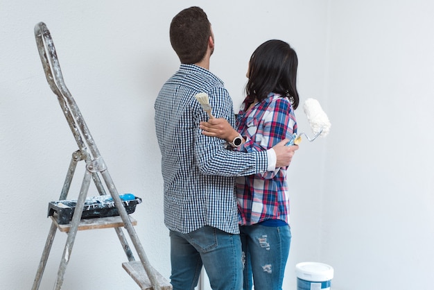 Paar mit Blick auf die Wandplanung und Entscheidungen zur Renovierung der Wohnung. Mann und Frau umarmen das Halten von Malwerkzeugen.