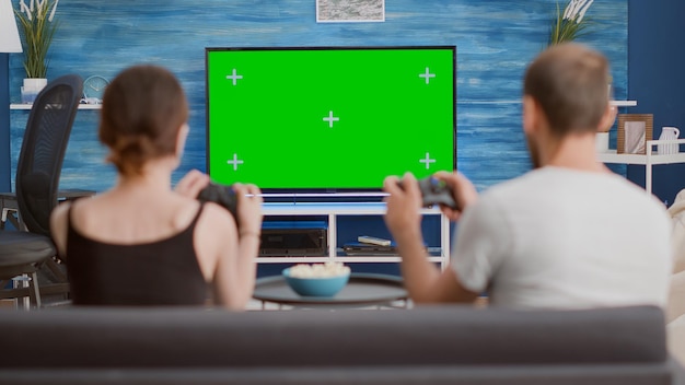 Paar mit Controllern, die Konsolen-Online-Spiele auf Greenscreen-Fernseher spielen, sitzen auf der Couch im modernen Wohnzimmer. Junge Frau und Freund verbringen Freizeit beim Spielen auf Chroma-Key-Mockup-Displays.