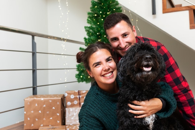 Paar mit schwarzem Pudelhund, der Kamera Weihnachtsbaum anschaut und im Hintergrund Urlaub präsent ist