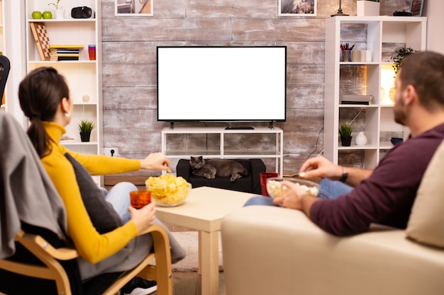 Paar schaut auf isolierten Fernsehbildschirm im gemütlichen Wohnzimmer, während es Essen zum Mitnehmen isst
