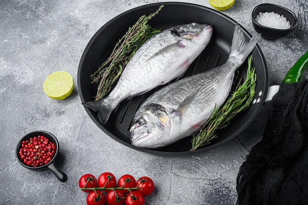 Paar Seabream oder Dorado roher Fisch auf Grillpfanne mit Zutaten auf grauweiß strukturiertem Hintergrund, Seitenansicht.