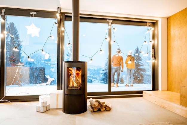 Paar steht im Winter draußen auf der Terrasse des Hauses mit Kamin. Innenansicht durch das Fenster