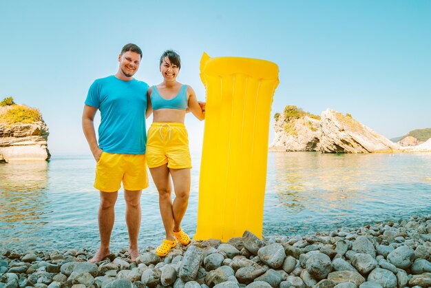 Paar steht mit gelber aufblasbarer Matratze am felsigen Sommerferienkonzept am Meer