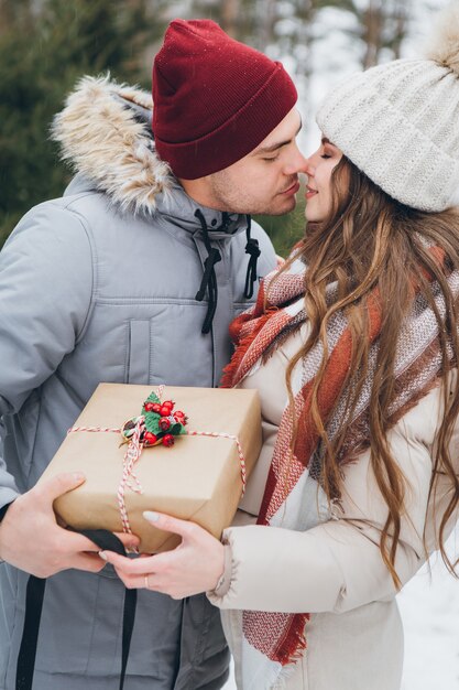 Paar umarmt und küsst sich in einem winterlichen Nadelwald, verschenkt sich Kisten mit Geschenken