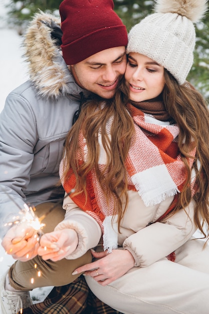 Paar umarmt und verbrennt Wunderkerzen in einem winterlichen Nadelwald, während es auf einem Schlitten sitzt