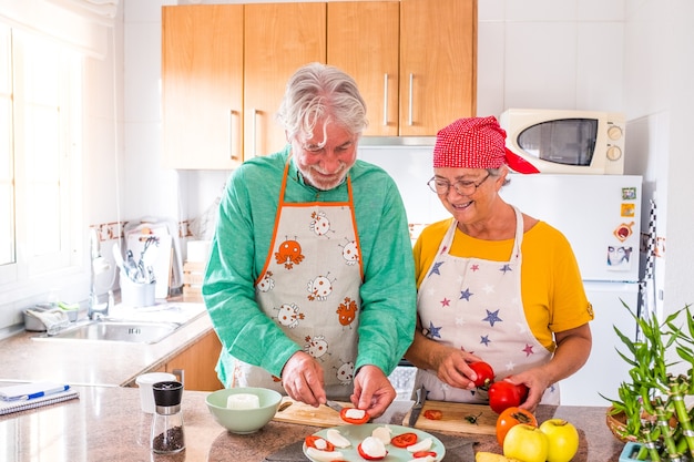 Paar von zwei glücklichen Senioren, die Spaß haben und zusammen in der Küche ihres Hauses kochen - gesundes Essen mit Tomaten zubereiten