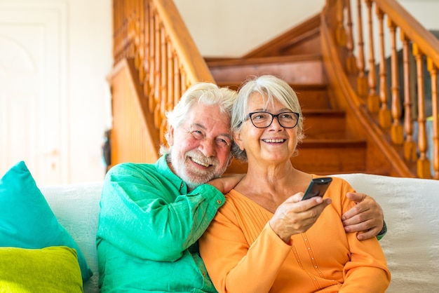 Paar von zwei glücklichen Senioren, die zu Hause auf dem Sofa sitzen, fernsehen und um die Fernbedienung des Fernsehers kämpfen