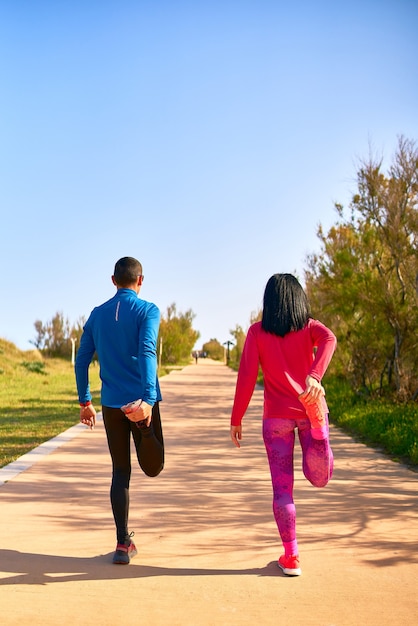 Paare, die ihre Beine vor dem Training ausstrecken. Die Frau trägt leuchtend rosa und lila Kleidung. Mann trägt blaues Hemd und schwarze lange Hose.