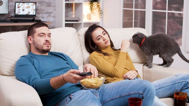 Paare, die sich entspannen, einen Film im Fernsehen ansehen und mit der Katze spielen. Mann mit TV-Fernbedienung und Chips essen.