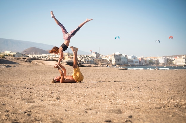 Paare von Erwachsenen in Beziehung oder Freundschaft, die zusammen Sport wie Acroyoga oder Yoga am Strand auf dem Sand treiben - allein und isoliert mit der Stadt im Hintergrund