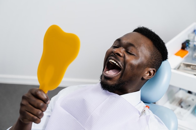 El paciente africano se mira en el espejo los dientes después de quitar la caries y rellenar los dientes Consulta con el dentista en la clínica dental El paciente afroamericano visita al dentista