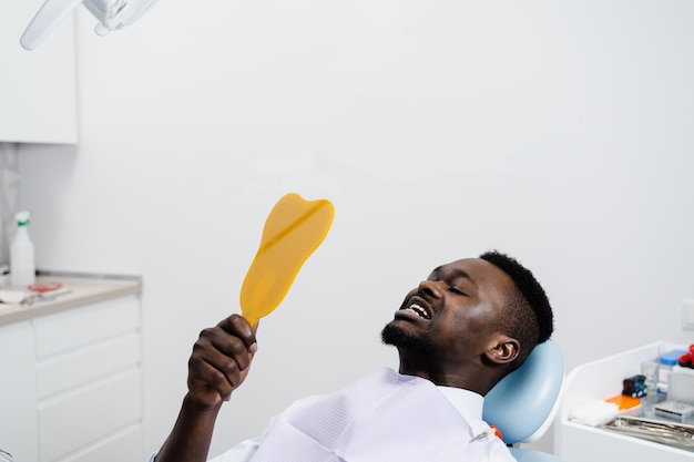 El paciente africano se mira en el espejo los dientes después de quitar la caries y rellenar los dientes Consulta con el dentista en la clínica dental El paciente afroamericano visita al dentista