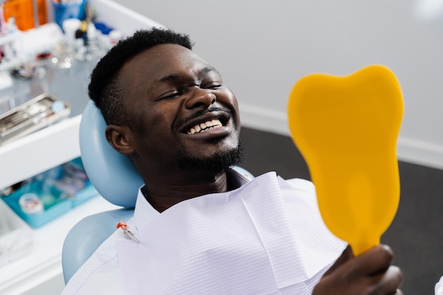 El paciente africano se mira en el espejo los dientes después de quitar la caries y rellenar los dientes Consulta con el dentista en la clínica dental El paciente afroamericano visita al dentista