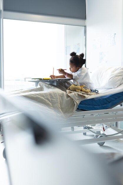 Foto paciente afro-americana deitada na cama a colorir no quarto do doente no hospital. hospital, infância, medicina e cuidados de saúde, inalterados.