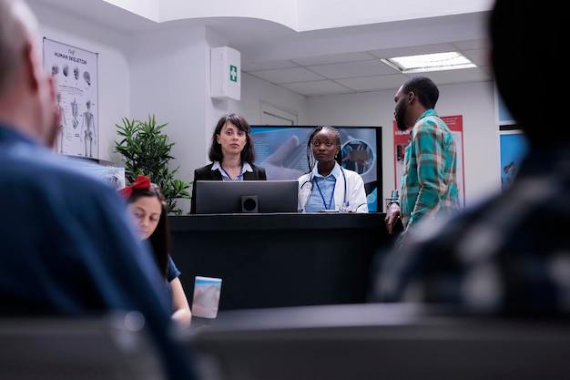 Paciente afro-americano se registrando para consulta médica na recepção da clínica privada conversando com médico e recepcionista. diversas pessoas conversando na sala de espera.