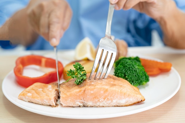 Paciente anciana asiática desayunando bistec de salmón con comida saludable vegetal mientras está sentada y hambrienta en la cama en el hospital