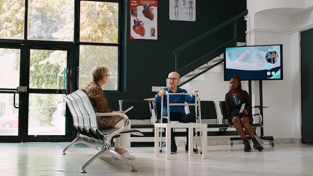 Paciente anciano hablando con personas en el vestíbulo de la sala de espera, usando un andador en la recepción del centro de salud. Hombre mayor con discapacidad esperando para asistir a un examen de consulta con un médico general.