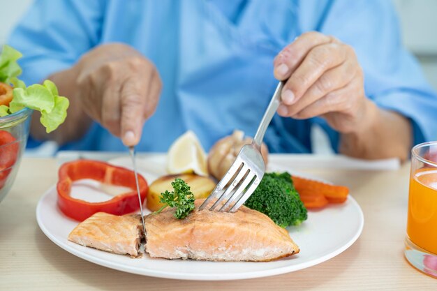 Paciente asiática mayor o anciana anciana comiendo filete de salmón desayuno con vegetales saludables mientras está sentada y hambrienta en la cama en el hospital