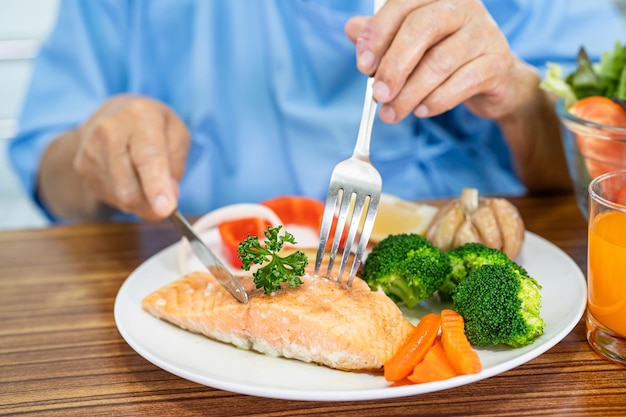 Paciente asiática mayor o anciana anciana comiendo filete de salmón desayuno con vegetales saludables mientras está sentada y hambrienta en la cama en el hospital