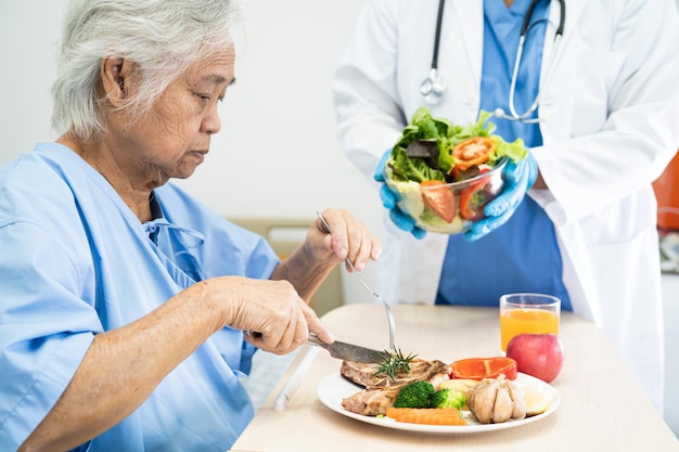 Paciente asiática mayor o anciana anciana desayunando y comiendo vegetales saludables con esperanza y feliz mientras está sentada y hambrienta en la cama en el hospital