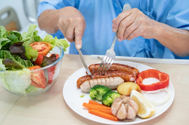 Paciente asiática mayor o anciana anciana desayunando y comiendo vegetales saludables con esperanza y feliz mientras está sentada y hambrienta en la cama en el hospital