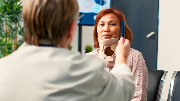 Paciente asiático com colar cervical fazendo consulta com especialista, fazendo exame de concussão para verificar a visão e danos cerebrais com luz. Comparecer à consulta de check-up com o médico. Tiro portátil.