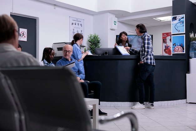 Paciente asiático entregando prancheta com formulário preenchido para recepcionista do hospital em pé na recepção. Homem atendendo a consulta médica em clínica privada movimentada com diversos pacientes.