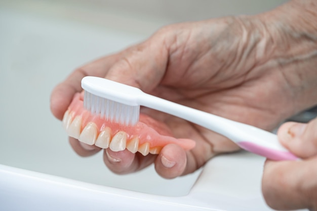 Paciente asiático mayor o anciana usa cepillo de dientes para limpiar la dentadura parcial de los dientes de reemplazo.