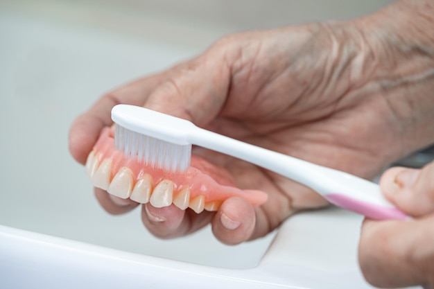 Paciente asiático sênior ou idosa usa escova de dentes para limpar próteses parciais de dentes substitutos.