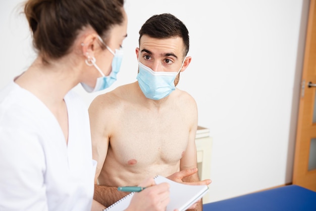 Foto paciente sin camisa charlando con el fisioterapeuta mientras usa una máscara facial médica durante la pandemia de coronavirus.