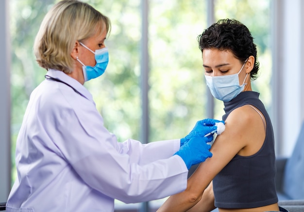 Paciente de mulher bonita senta-se à espera de covid 19 vacinando com agulha de seringa após médico feminino branco em jaleco branco com máscara facial e luvas de borracha usar ombro de limpeza de algodão de álcool.