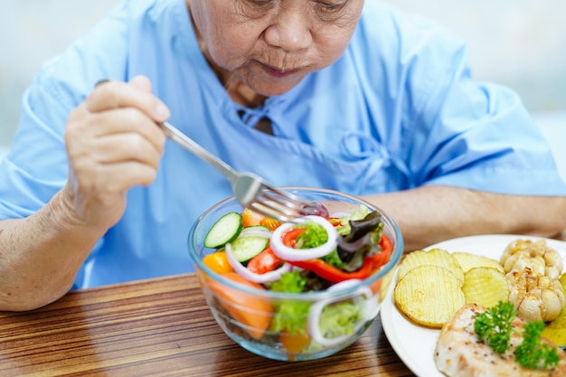 Paciente de mulher idosa asiática sênior ou idosa comendo alimentos saudáveis de vegetais de café da manhã com esperança e feliz enquanto está sentado e com fome na cama no hospital.