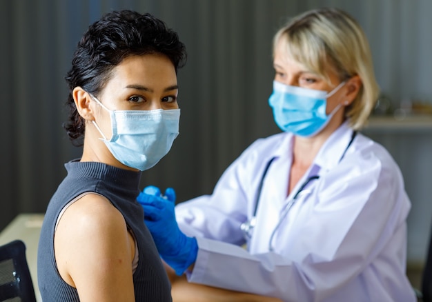 Paciente do sexo feminino de cabelo preto curto bonito usa máscara facial sentar olhar para a câmera enquanto médico caucasiano em jaleco branco com estetoscópio injetando vacina em seu ombro no fundo desfocado.