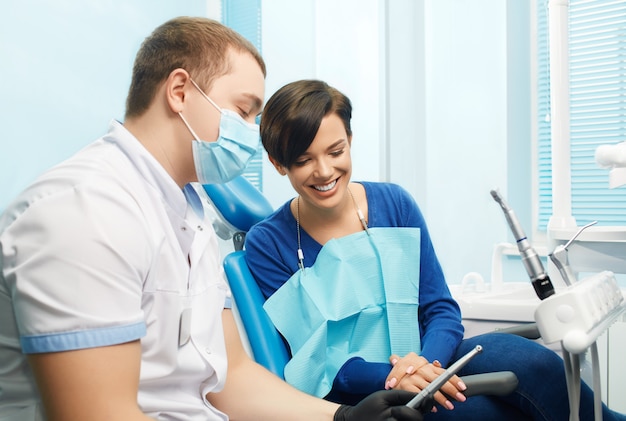 Paciente do sexo feminino jovem visitar consultório dentista. Bela mulher sentada na cadeira odontológica. Dentista, mostrando o equipamento dental. Clinica odontológica. Estomatologia