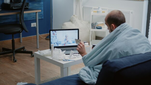 Paciente enfermo hablando en videoconferencia para telesalud usando una computadora portátil. Hombre sosteniendo una tableta de cápsula, preguntando al médico sobre medicamentos recetados y tratamiento mientras usa una conferencia remota en línea