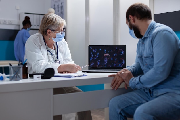 Paciente y especialista con mascarilla mirando la ilustración del virus en la computadora portátil