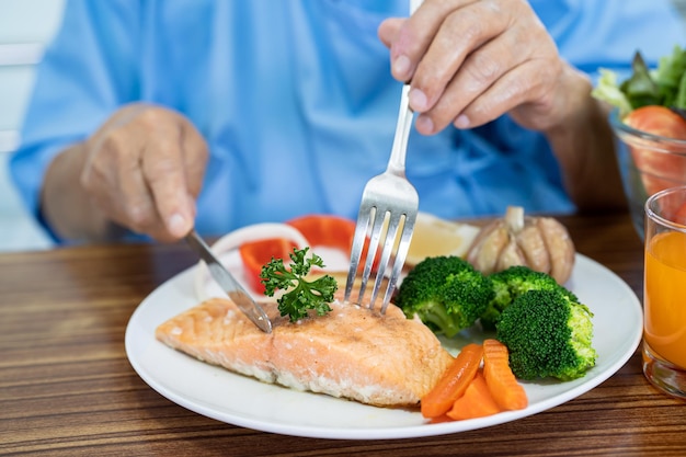 Paciente idosa asiática comendo bife de salmão no café da manhã com comida saudável vegetal enquanto está sentada e com fome na cama no hospital