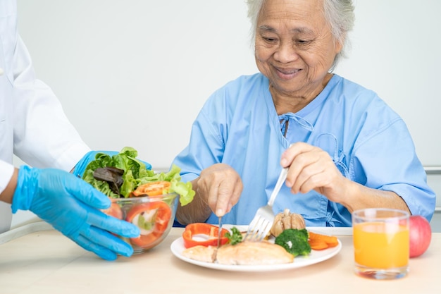Paciente idosa asiática ou idosa tomando café da manhã de bife de salmão com comida saudável vegetal enquanto está sentado e com fome na cama no hospital